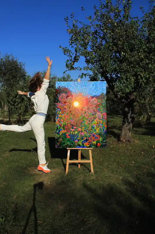 A person in white athletic wear strikes a dynamic, arms-raised pose next to an easel outdoors. The canvas on the easel…