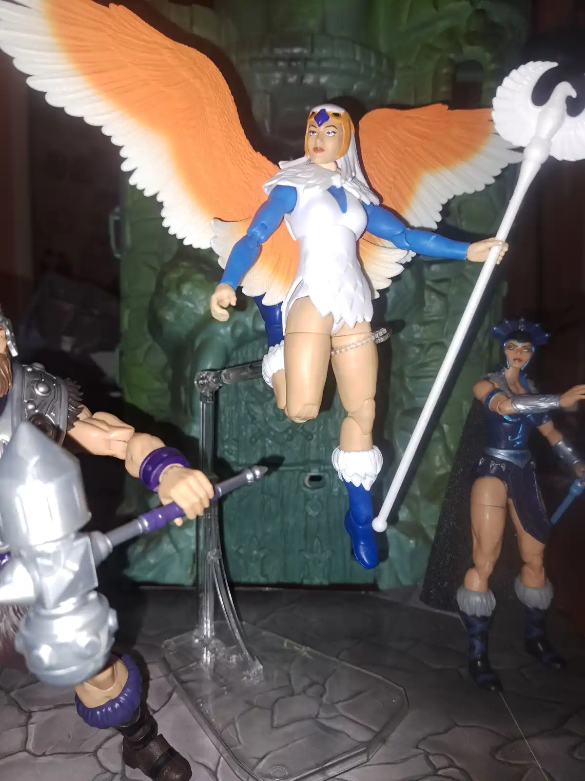 A toy action figure of a female character with large orange and white wings, wearing a white and blue costume, is suspended…