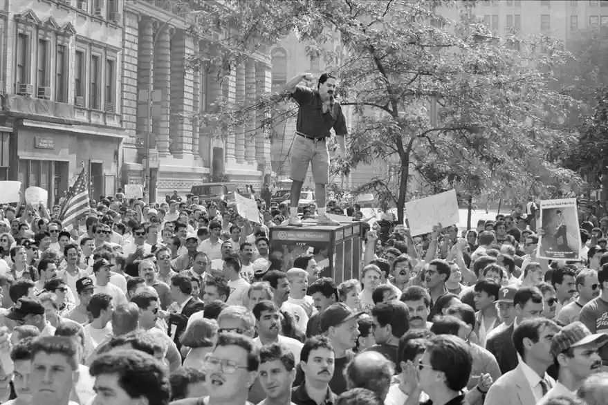 a black-and-white photo shows a man with a mustache standing atop a telephone booth, arms raised, addressing a large crowd…