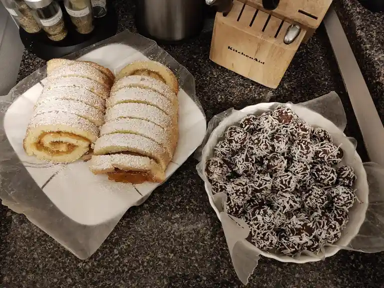 A kitchen counter displays two baked goods: a sliced cake roll dusted with powdered sugar on a white plate, and a bowl of…