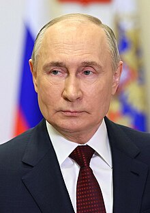 A formal portrait of Vladimir Putin, dressed in a dark suit and red patterned tie, with a stern expression. Behind him, the…