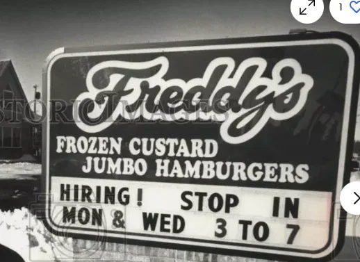 A black-and-white photo of a vintage sign for 'Freddy's Frozen Custard Jumbo Hamburgers' advertising a hiring event. The…