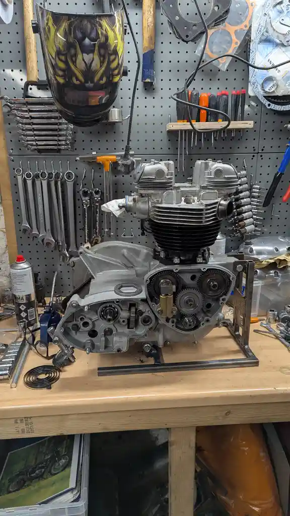 A partially disassembled motorcycle engine sits on a workbench in a cluttered garage, surrounded by tools like wrenches…