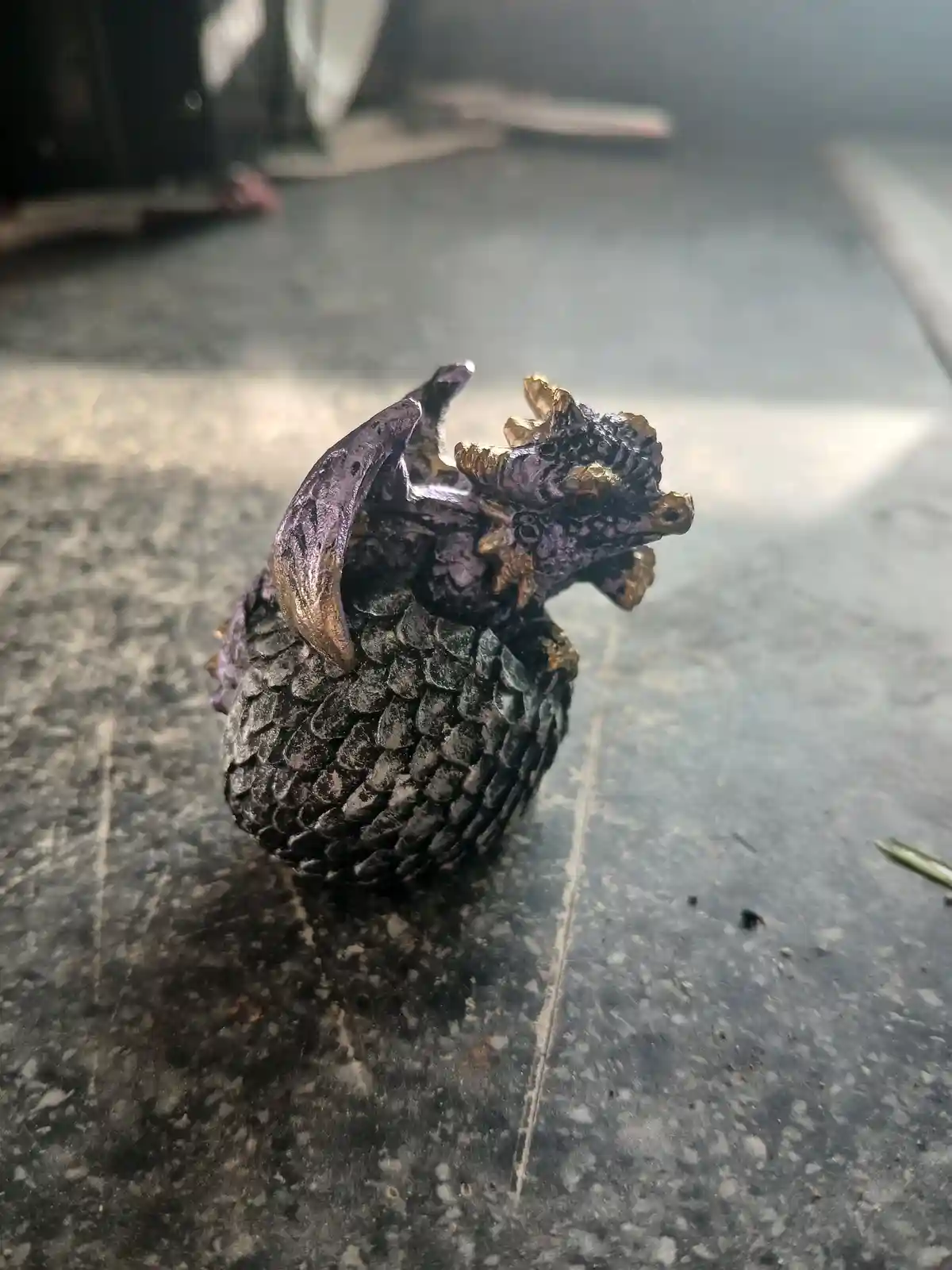 A small, intricately detailed figurine of a dragon with purple and gold accents is sitting on a dark, speckled floor. The…