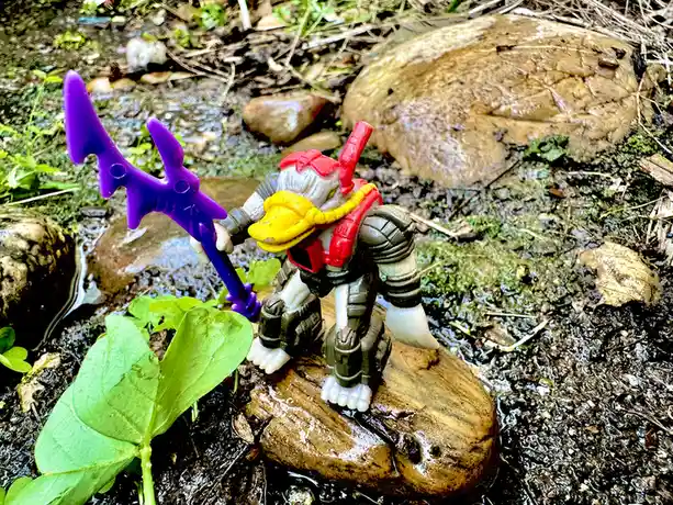 A small plastic action figure with a duck-like head, wearing red and yellow armor and holding a purple axe, stands on a wet…