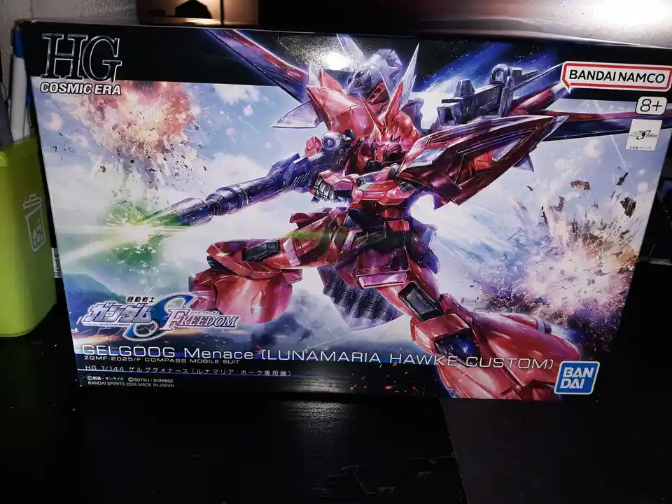 A photo of a Bandai HG Cosmic Era Gundam Seed Freedom model box featuring the red and purple Gelgoog Menace mobile suit…