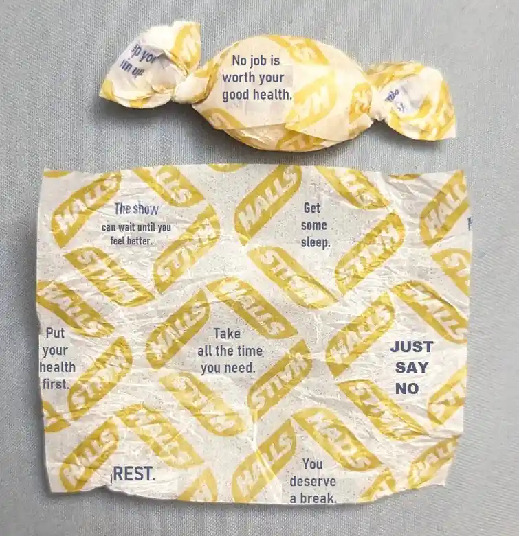 A crumpled Halls cough drop wrapper and a wrapped cough drop are laid out on a gray surface. The wrapper has multiple…