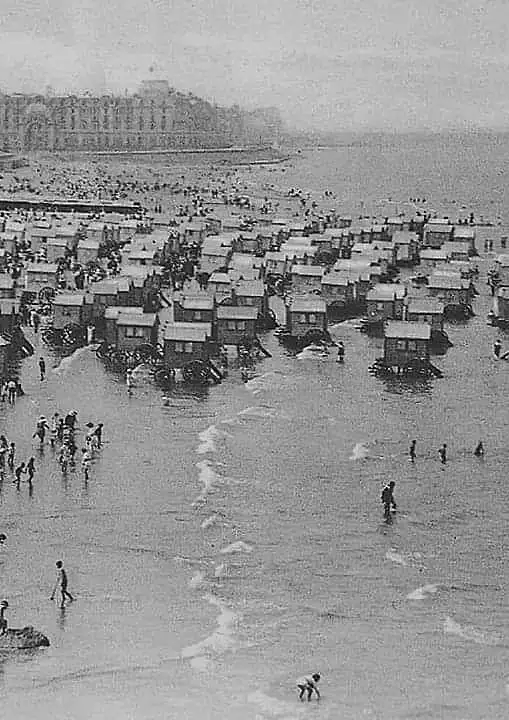 a black-and-white historical photo shows a crowded beach filled with numerous small, wooden beach huts built on stilts in…