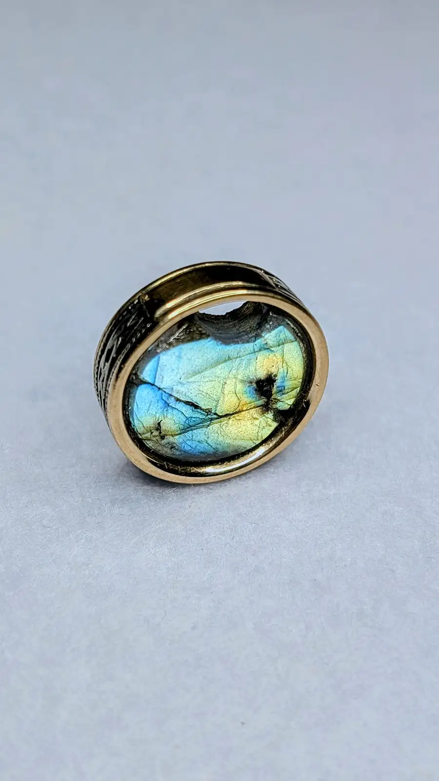 A close-up photo of a ring with a large, oval labradorite gemstone set in a gold-toned band, resting on a plain light gray…