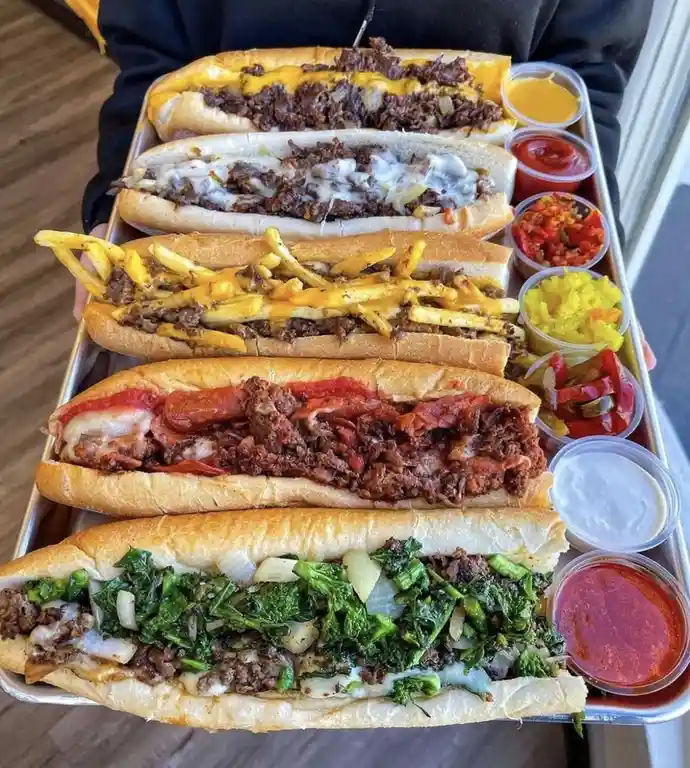 A person holds a metal tray with five large, generously loaded submarine sandwiches stacked vertically, each with different…