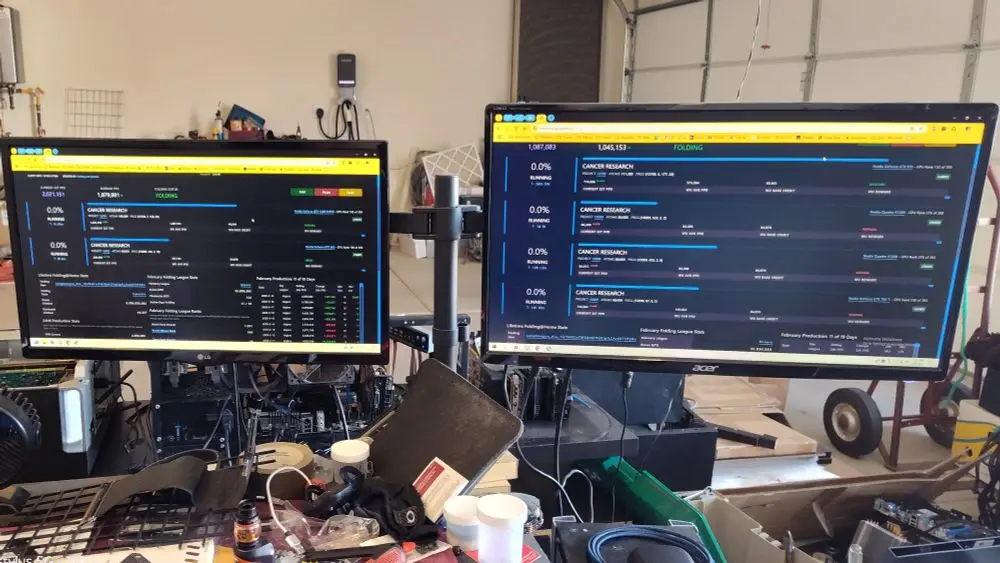 Two computer monitors in a cluttered garage display the BOINC Folding@home interface, showing 'CANCER RESEARCH' projects…