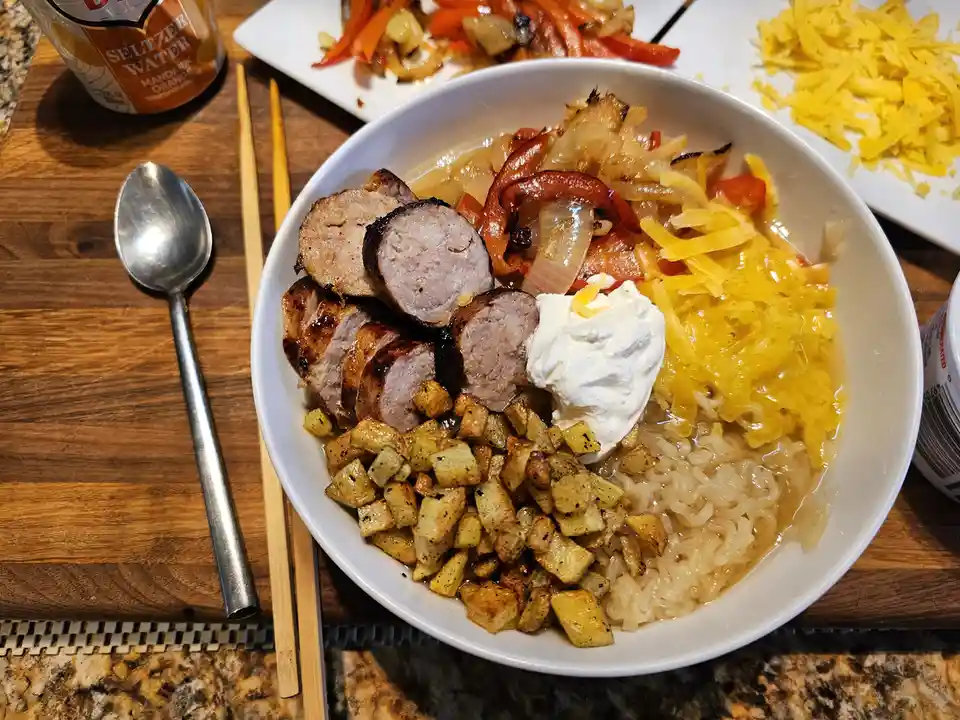 A white bowl filled with ramen noodles, sliced sausage, roasted potatoes, sautéed peppers and onions, shredded cheese, and a…