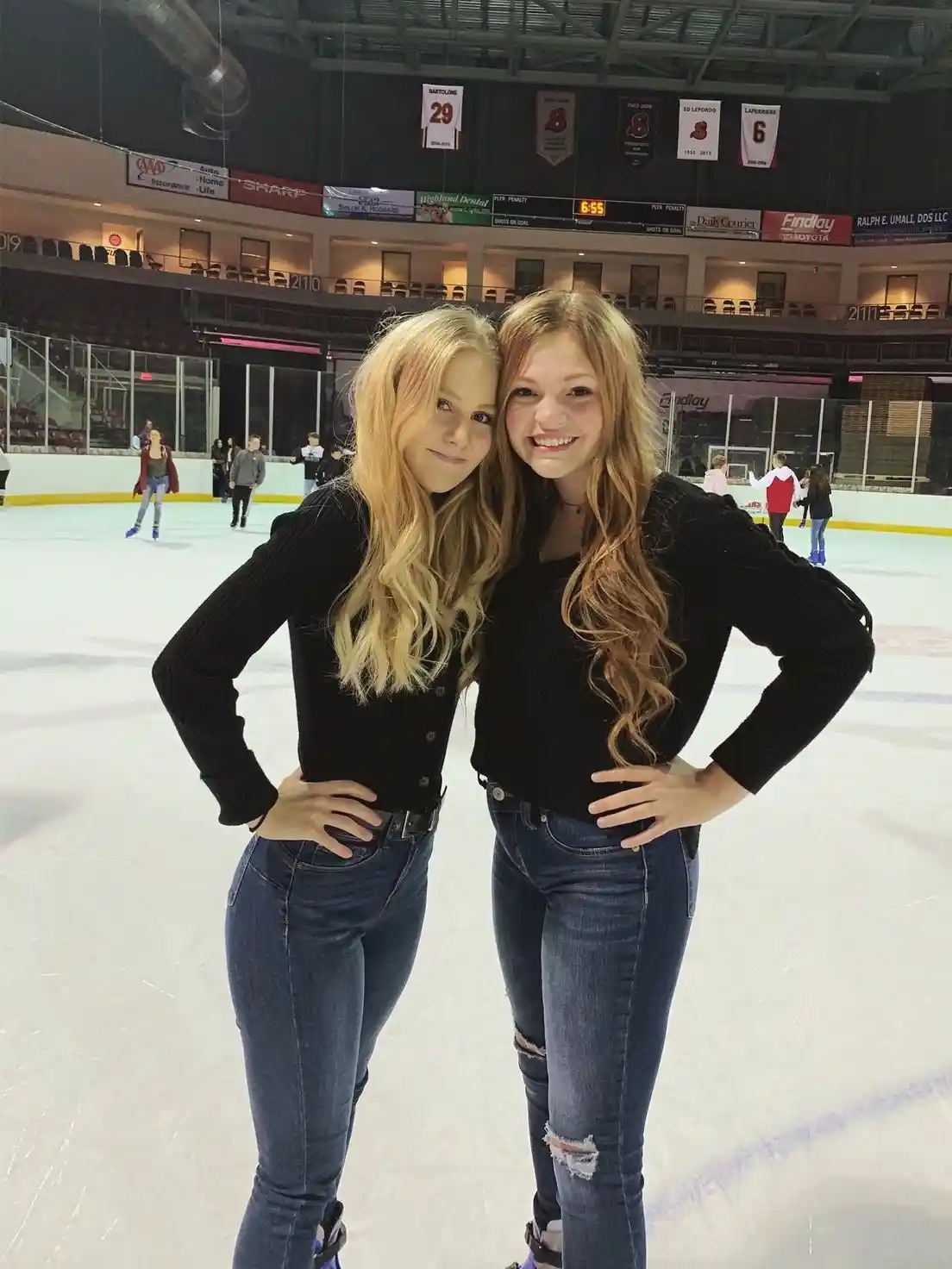 Two young women in matching black sweaters and blue jeans pose confidently with hands on hips on an ice rink, smiling at the…