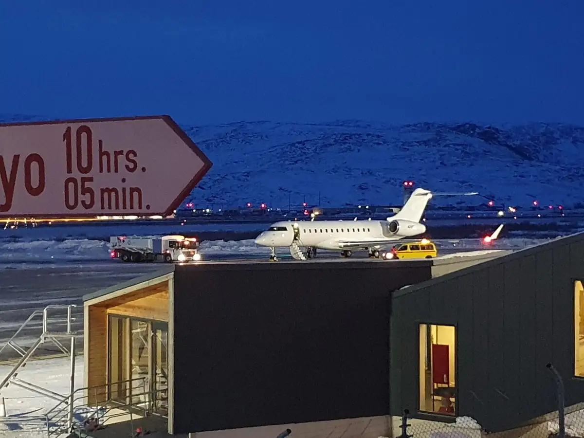 A white private jet sits on a snowy airport tarmac at dusk, with a large directional sign partially visible that reads 'yo…