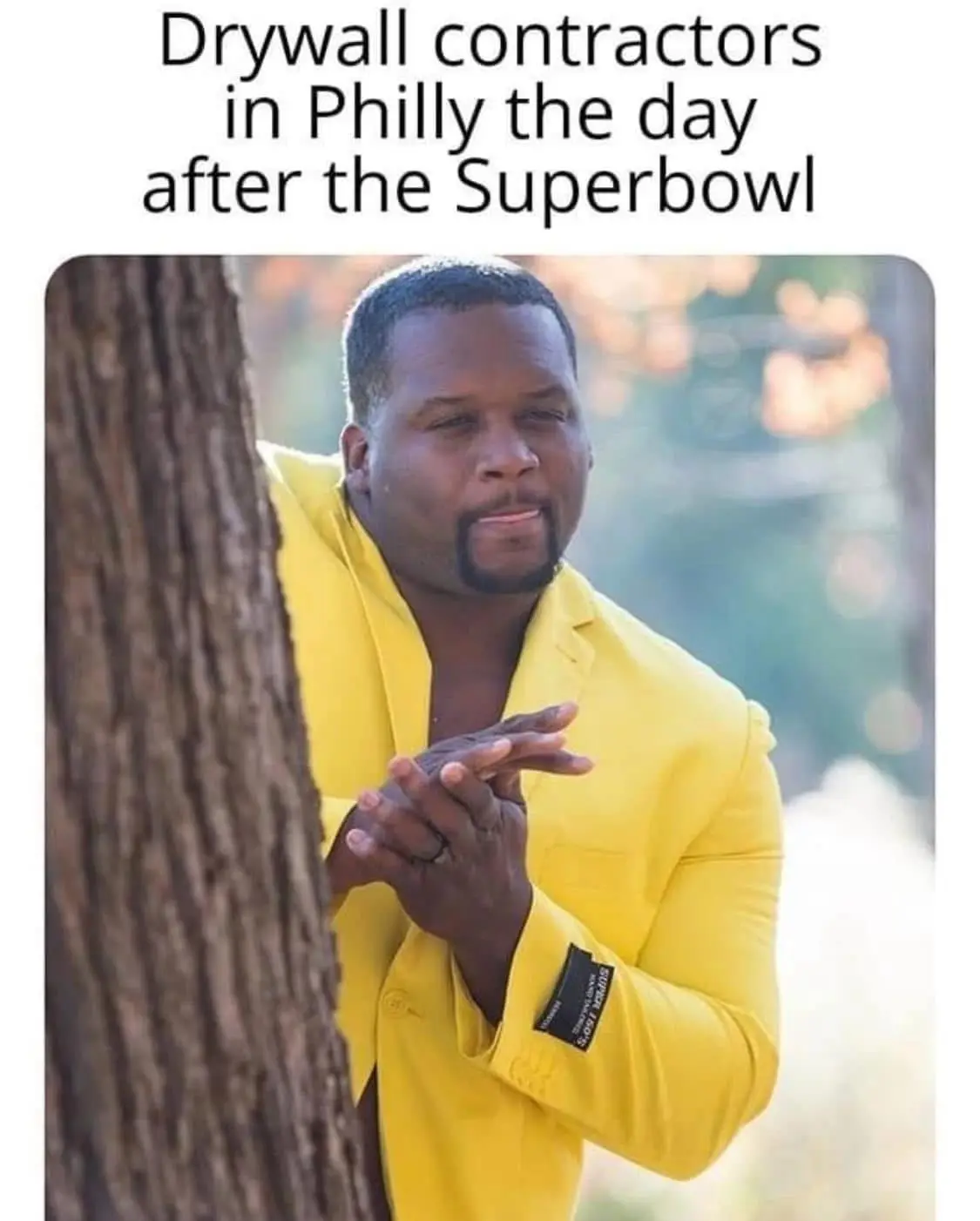 A man in a bright yellow blazer peeks out from behind a tree trunk with a smug, self-satisfied expression, hands clasped…