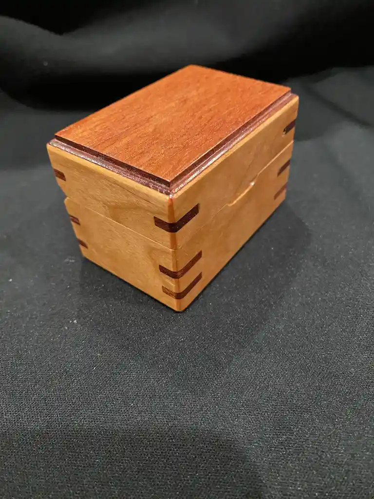 A small, handcrafted wooden box with a light-colored body and a darker reddish-brown lid sits on a black textured surface…