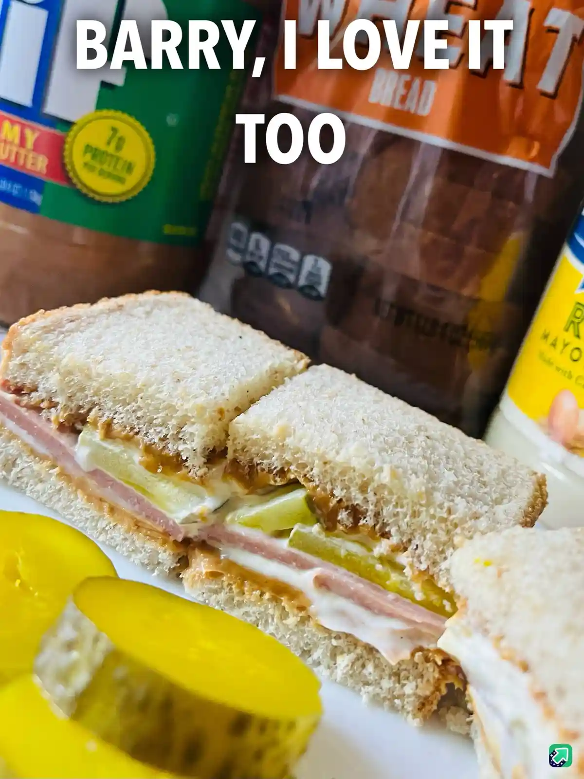 A close-up photo of a sandwich cut in half, made with white bread, ham, pickles, and a creamy spread, sits on a plate with…