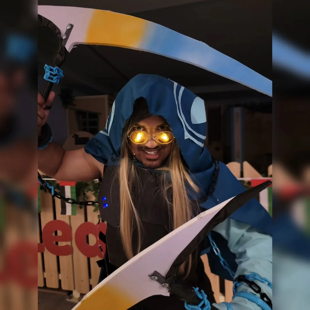 A person in a blue hooded costume with glowing yellow goggles and long blonde wig holds two large, colorful, curved swords…