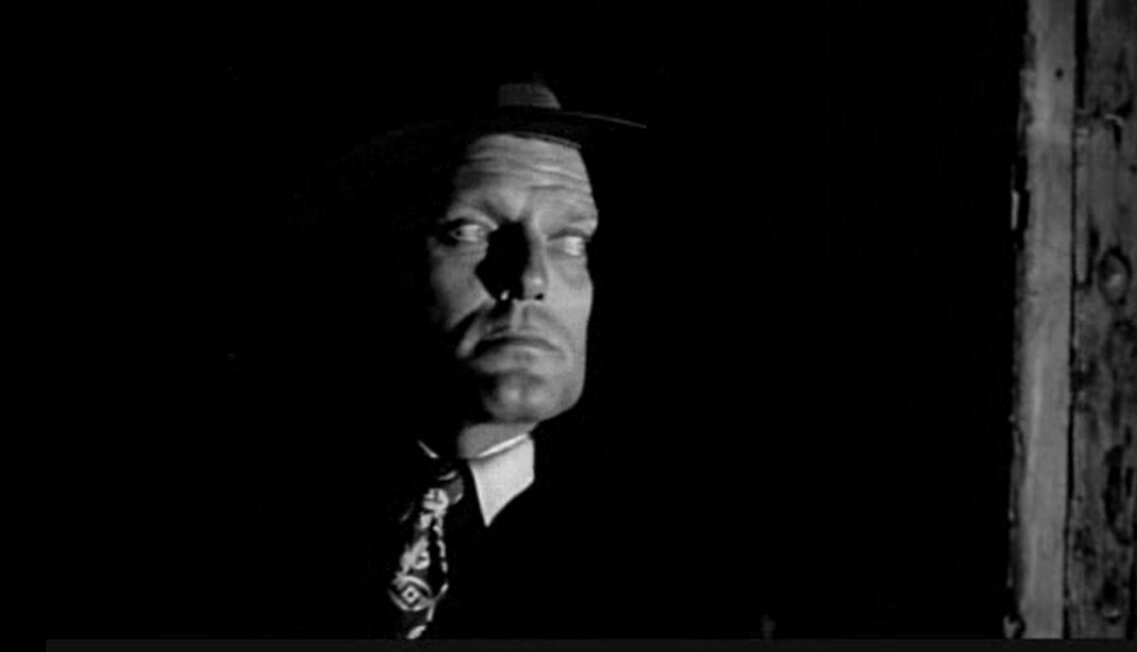A black-and-white still shows a man in a hat and tie, partially obscured by shadow, with a stern, unimpressed expression…