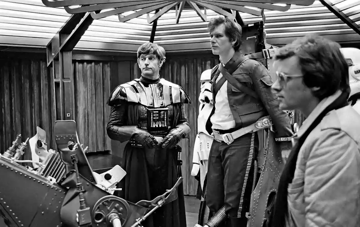 A black-and-white behind-the-scenes photo from Star Wars shows three actors in costume standing near a spaceship cockpit…