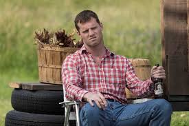 a man in a red plaid shirt and jeans sits on a chair outdoors, holding a beer bottle, with a basket of dried plants and…