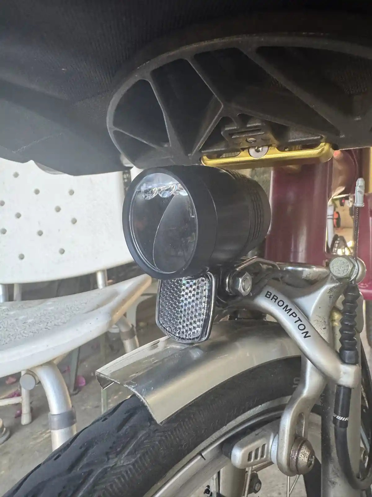 A close-up photo shows a black cylindrical LED light mounted on the front of a Brompton bicycle, positioned under the seat…