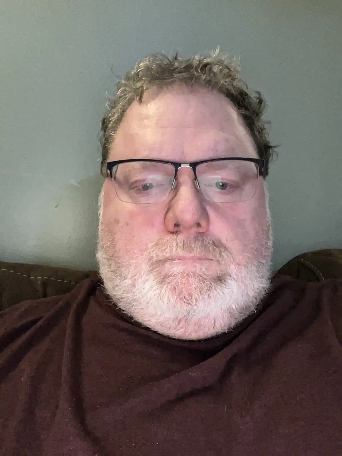 A close-up photo of a man with glasses, a white beard, and curly hair, wearing a brown shirt and sitting on a couch against…