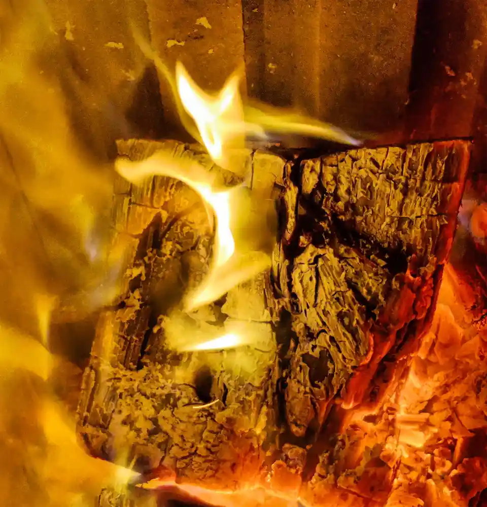 A close-up photo of a burning log in a fireplace, with bright yellow-orange flames licking upwards from the charred wood…
