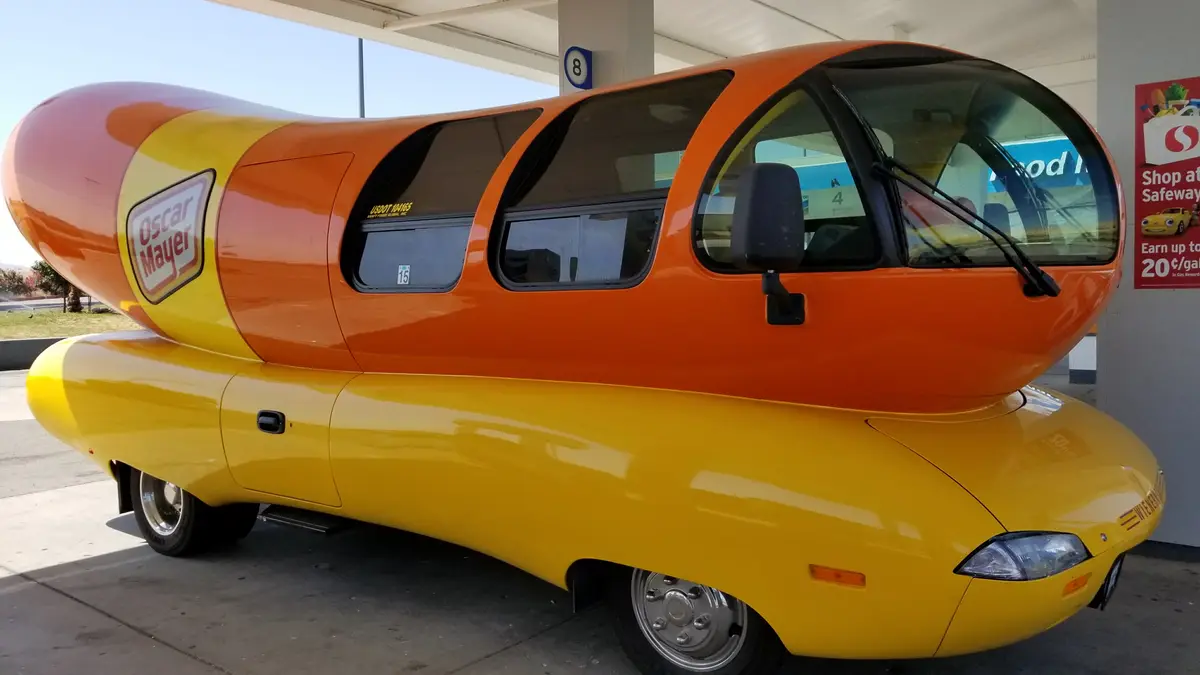 A large, orange-and-yellow vehicle shaped like a hot dog with the Oscar Mayer logo is parked at a gas station pump. The car…