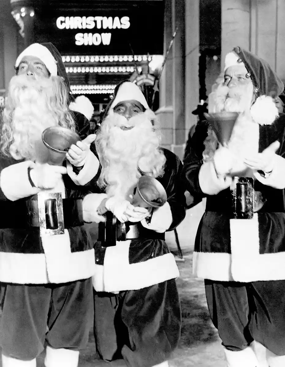 Three men dressed as Santa Claus stand in front of a theater marquee that reads 'CHRISTMAS SHOW'. Each Santa holds a small…
