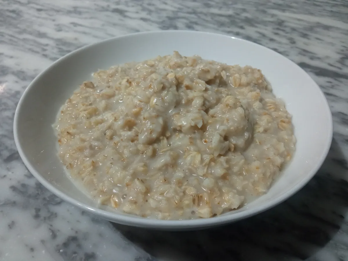 A close-up photo shows a white bowl filled with plain, creamy oatmeal resting on a gray and white marble countertop. The…