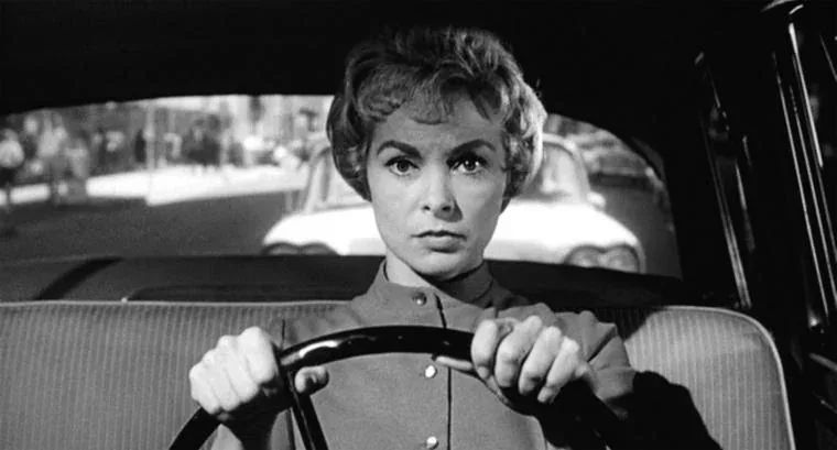 A black-and-white still of a woman with short, styled hair gripping a car steering wheel with both hands, looking intensely…