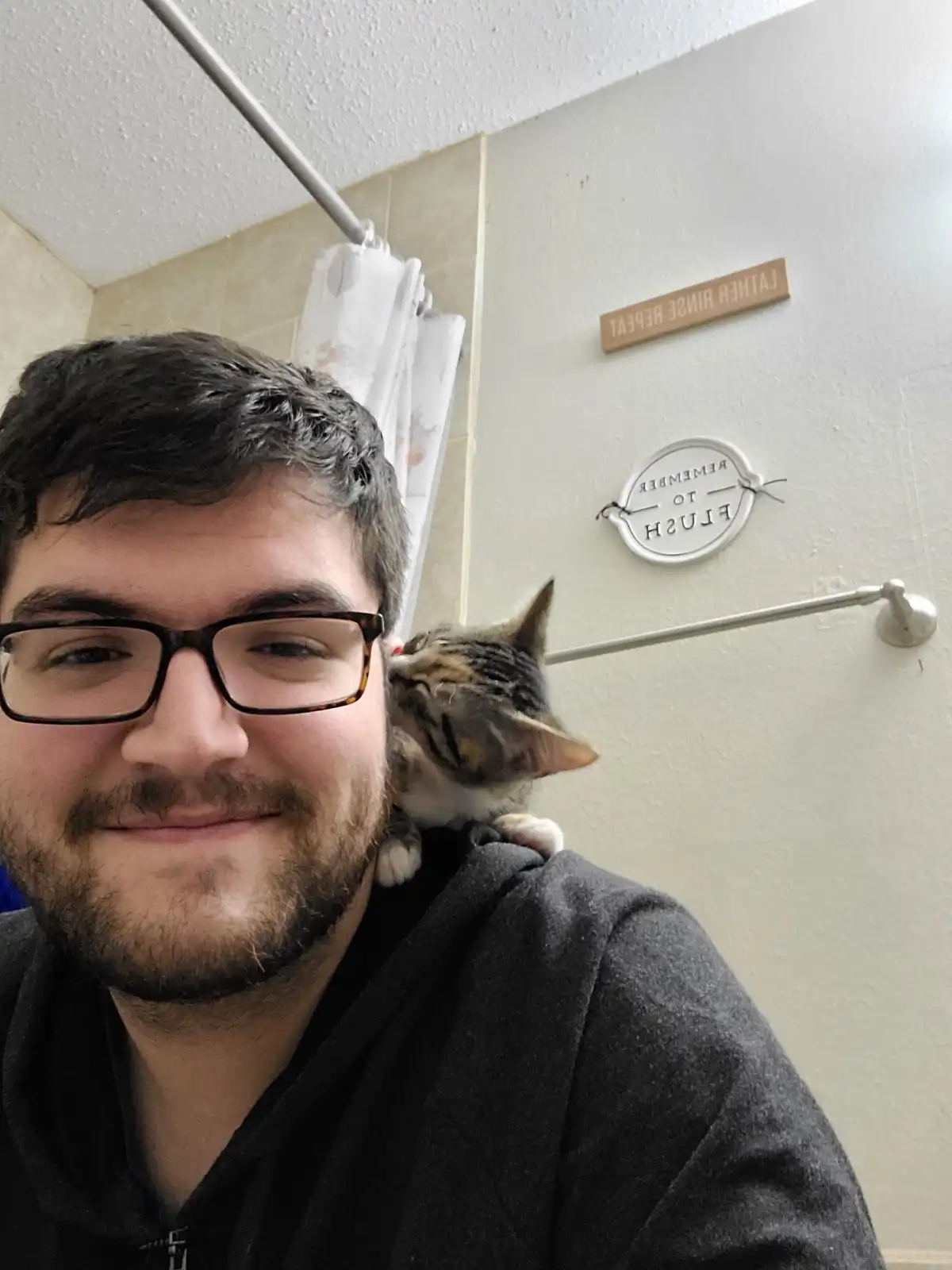 A man with glasses and a beard smiles at the camera while a small tabby cat nuzzles his ear from behind his shoulder in a…
