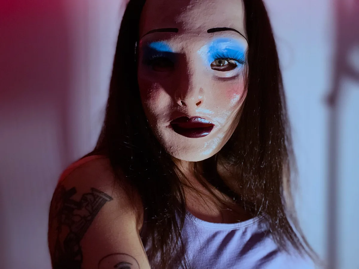 A person with long dark hair and a visible tattoo on their shoulder wears heavy, doll-like makeup including thick black…