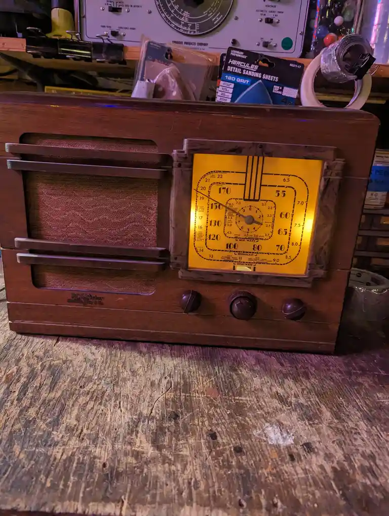 A vintage wooden RCA Victor radio with a glowing yellow dial sits on a worn wooden table, surrounded by workshop clutter…