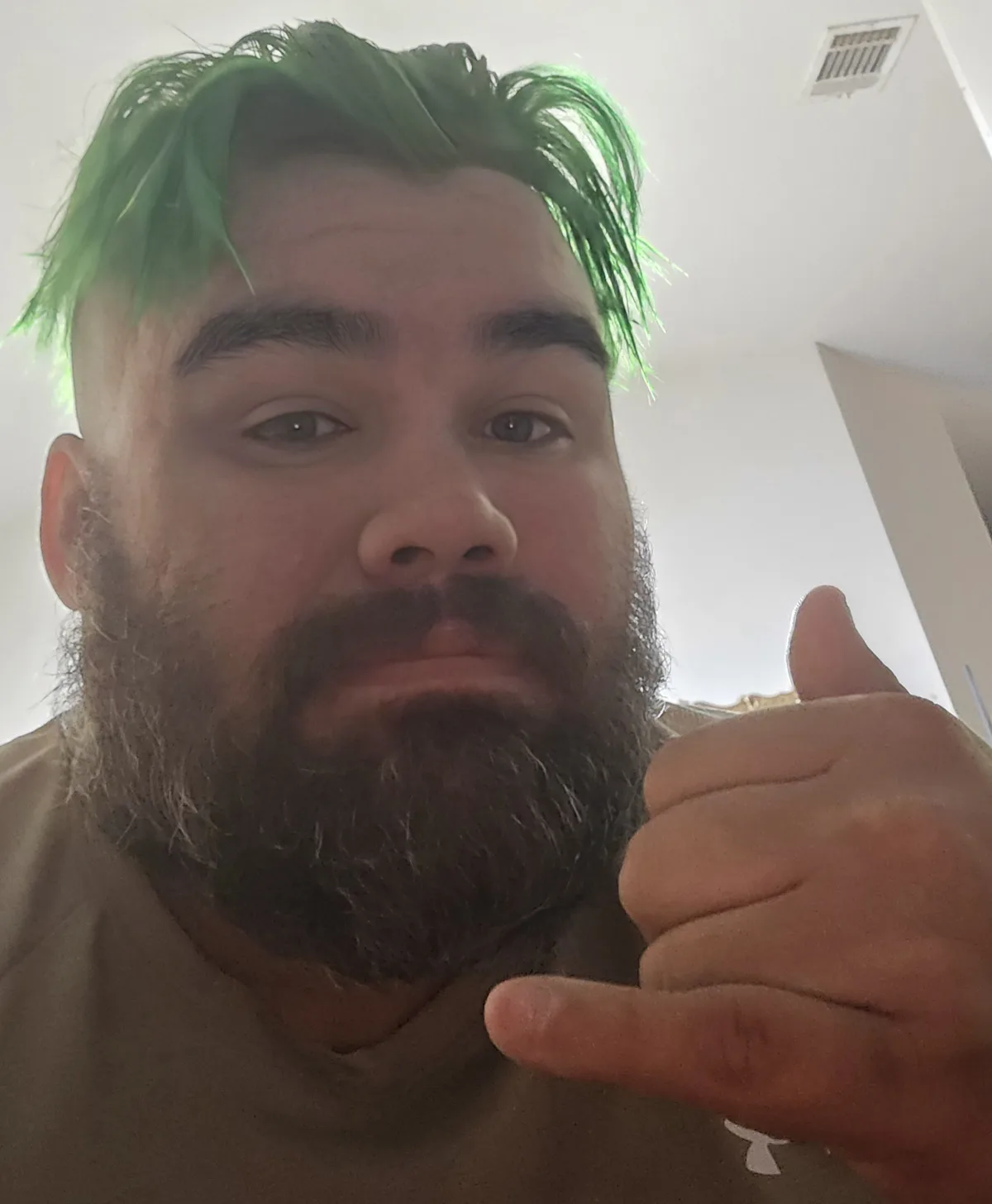 A man with bright green hair and a full beard looks directly at the camera while making a 'call me' hand gesture. The…