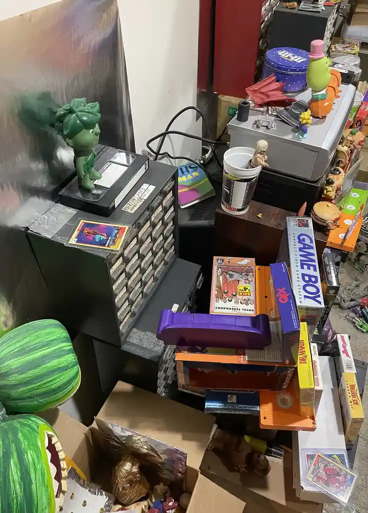 A cluttered, chaotic pile of vintage video game consoles, cartridges, VHS tapes, action figures, and collectibles fills the…
