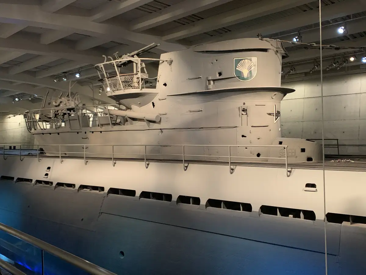 A large, gray submarine is displayed indoors, likely in a museum, with its conning tower and deck visible. The conning tower…