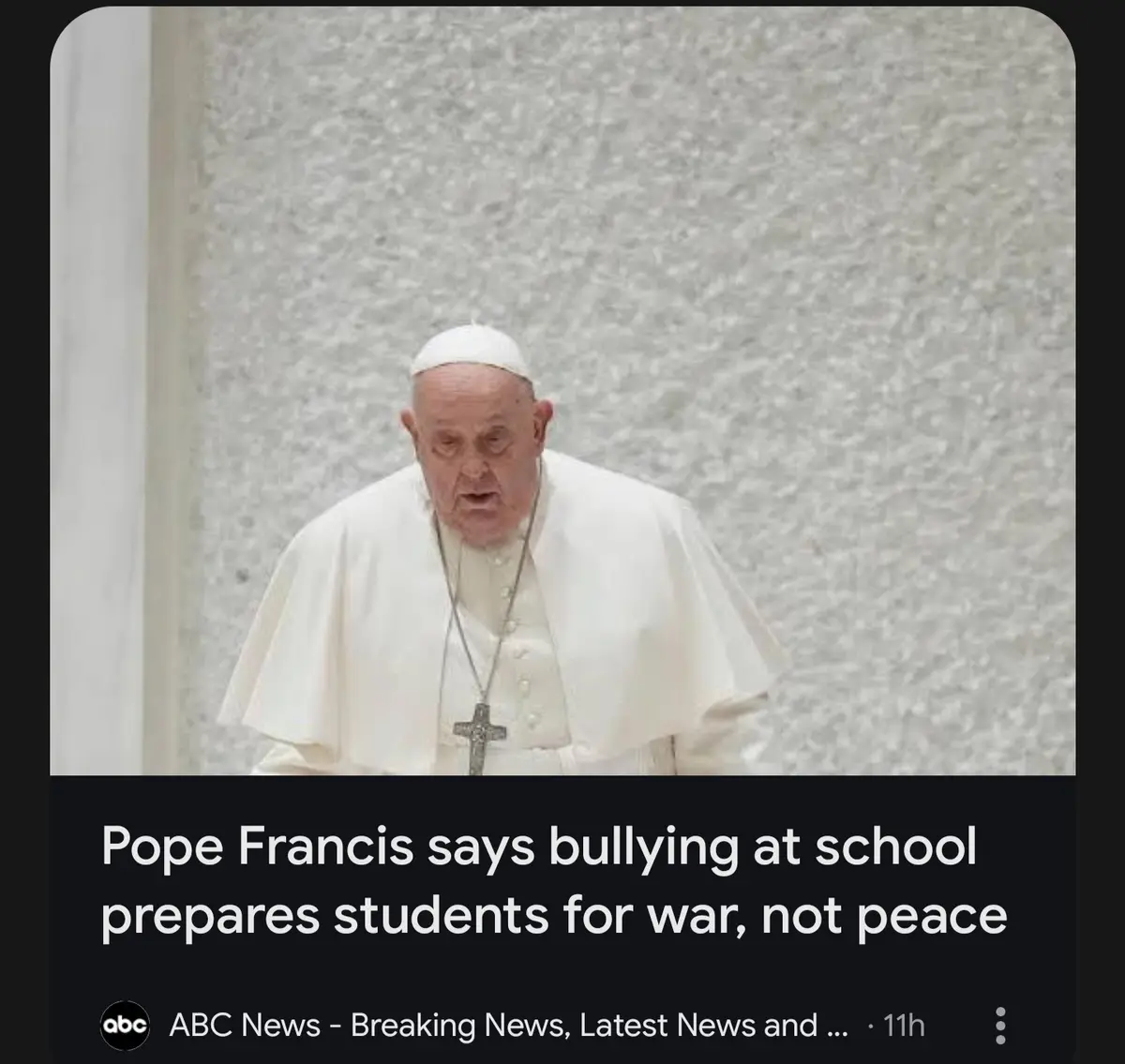 The image shows Pope Francis in his white papal attire, looking down with a serious expression against a textured gray wall…