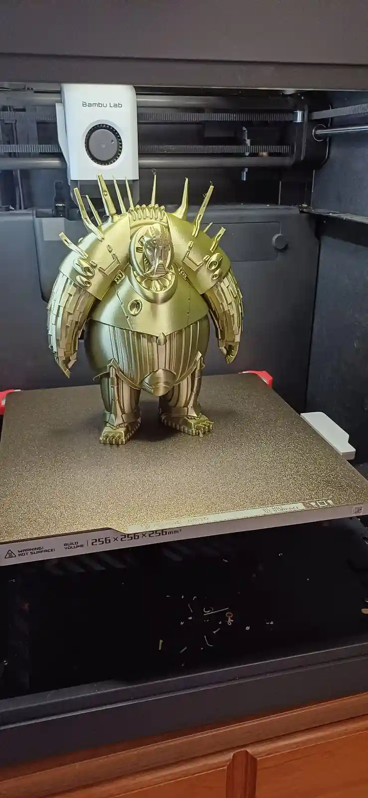 A golden, intricately detailed 3d-printed figurine of a bulky, armored character with spikes and a helmet stands on the…