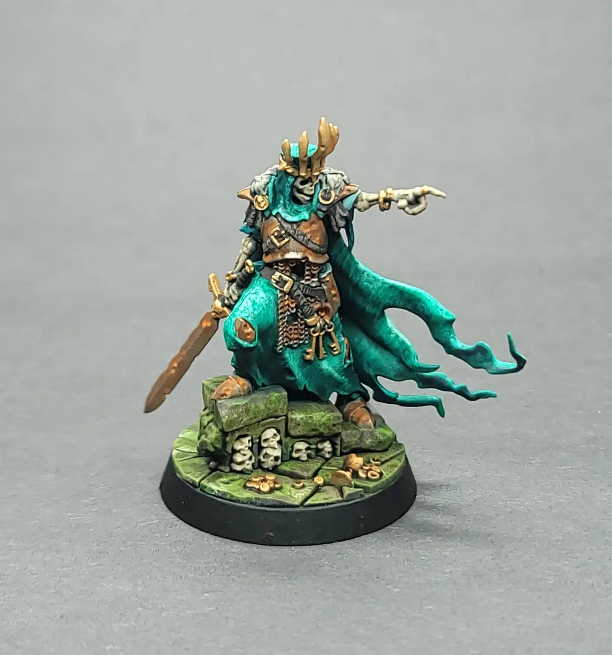 A painted miniature of a skeletal, teal-robed fantasy warrior with antler-like headgear, pointing forward while holding a…