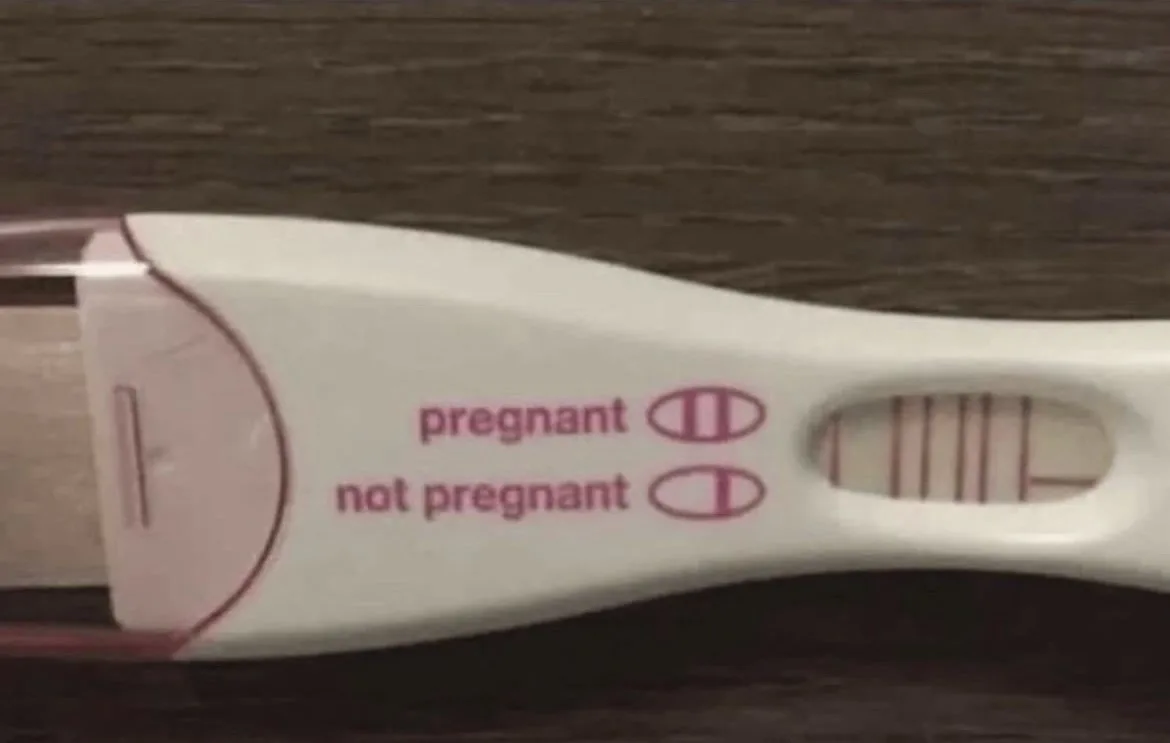 A close-up photo of a white and pink pregnancy test lying on a dark wooden surface. The test has printed labels 'pregnant'…