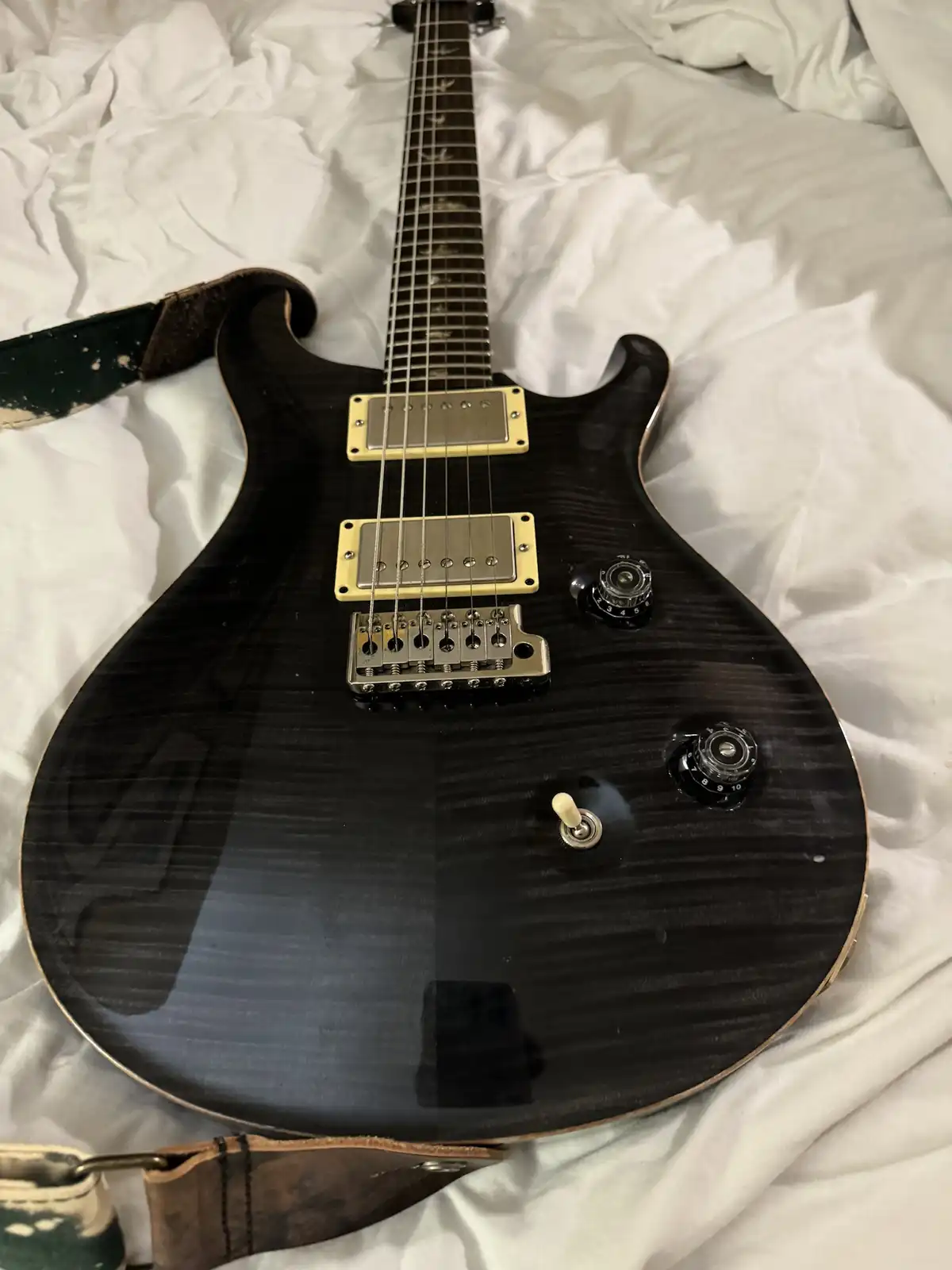 A black electric guitar with gold hardware and a worn leather strap lies on a rumpled white bedspread. The overlaid text…