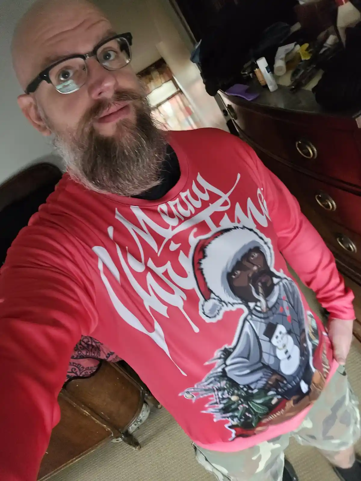 A bald man with glasses and a full beard takes a selfie wearing a red long-sleeved shirt with a graphic of a man in a Santa…