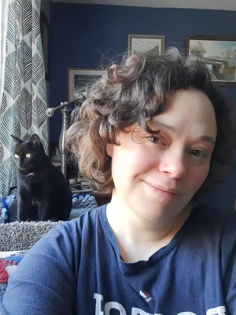 A person with curly brown hair and a slight smile looks at the camera while a black cat with glowing eyes sits behind them…