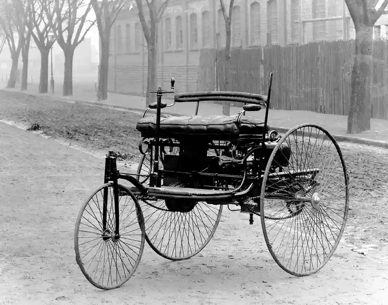 A black-and-white historical photograph shows an early 19th-century automobile, likely the Benz Patent-Motorwagen, parked on…