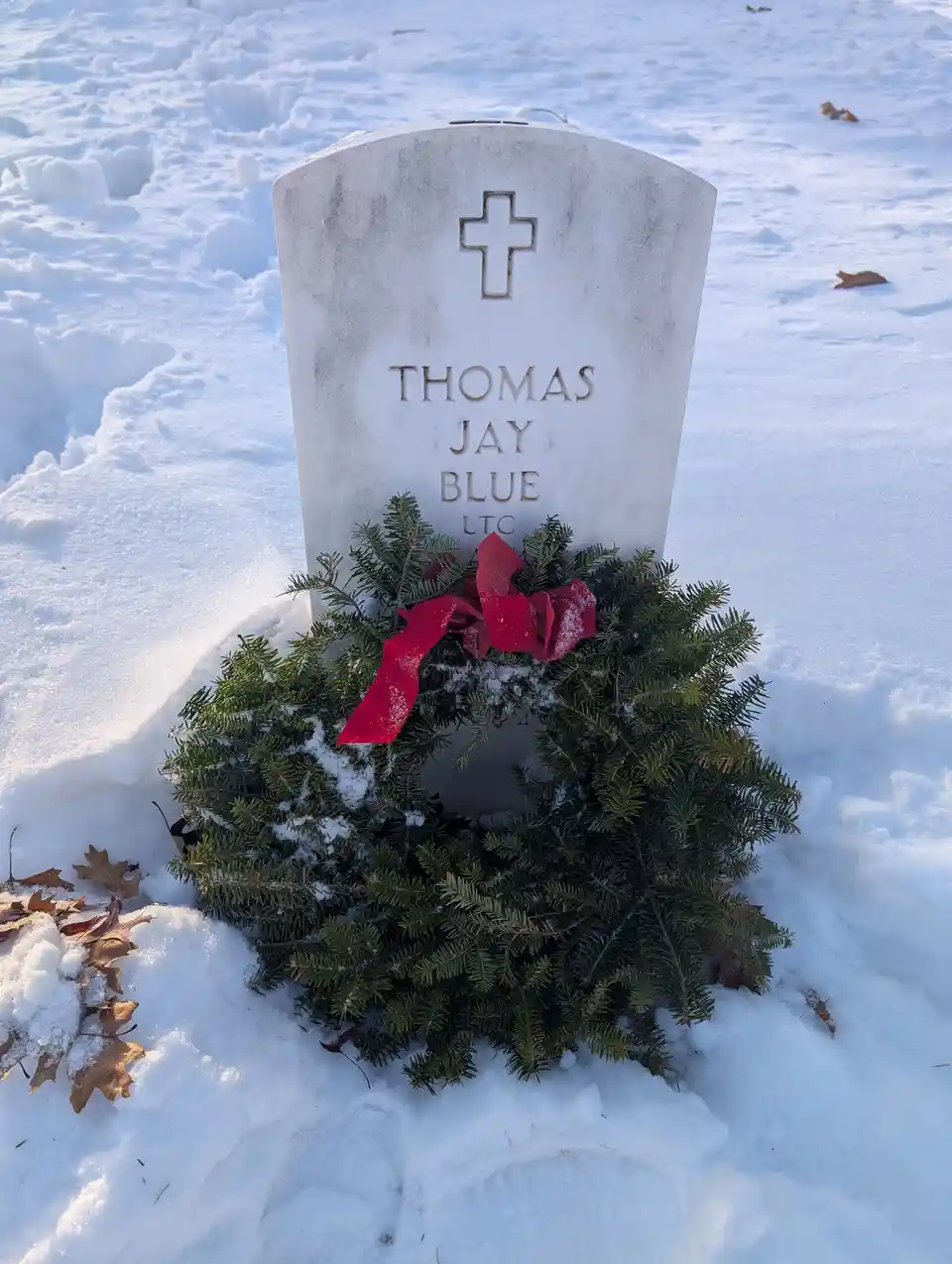 A white military gravestone in a snowy cemetery, engraved with 'THOMAS JAY BLUE LTC' and topped with a cross, has a green…