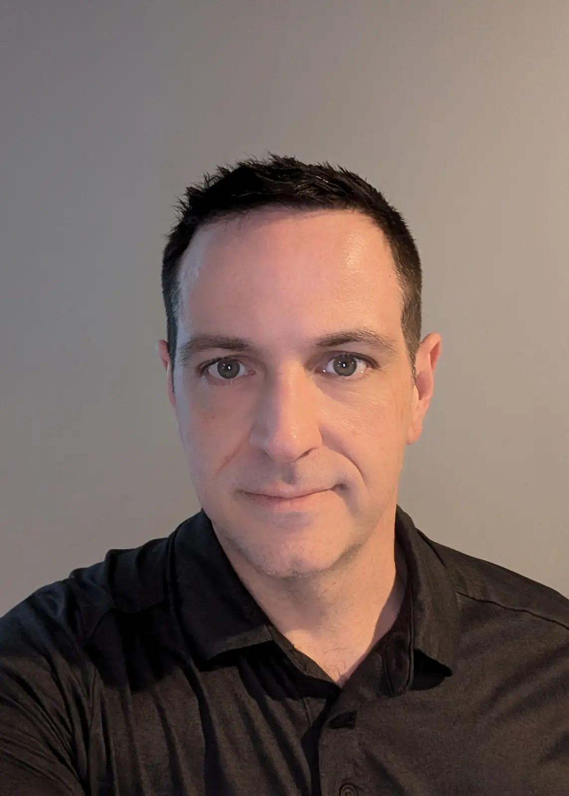 A close-up selfie of a man with short dark hair and green eyes, wearing a dark collared shirt, looking directly at the…