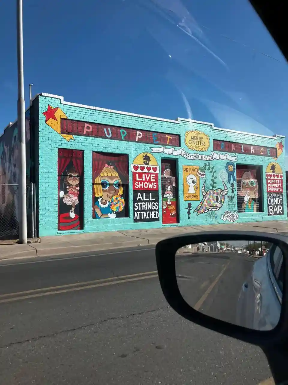 A photo taken from inside a car shows a brightly painted turquoise building with a mural of cartoon puppets in windows and…