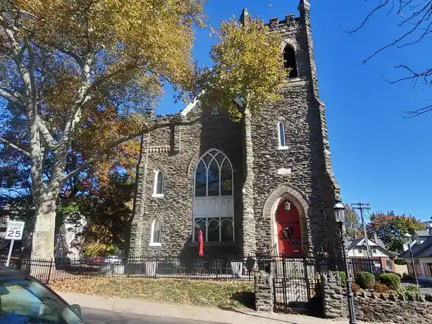 A stone church with a red door and Gothic windows stands under a clear blue sky, surrounded by autumn trees with yellow and…